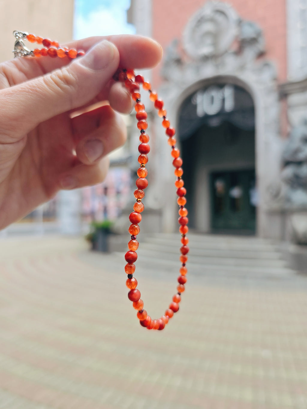 Hand holding a red, orange beaded necklace with a blurred background of a art deco building entrance.